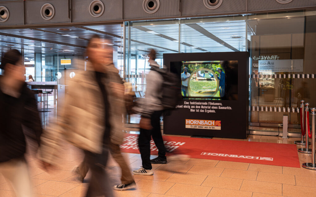 Reisende passieren am Hamburg Airport den Hornbach-Aufbau, auf dessen Display der Kurzfilm aus der Baumarkt-Kamera läuft. (Foto: Hamburg Airport Media)